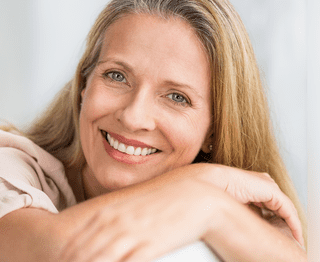 Smiling woman with long blonde hair resting head on arms against white background, dental clinic portrait.