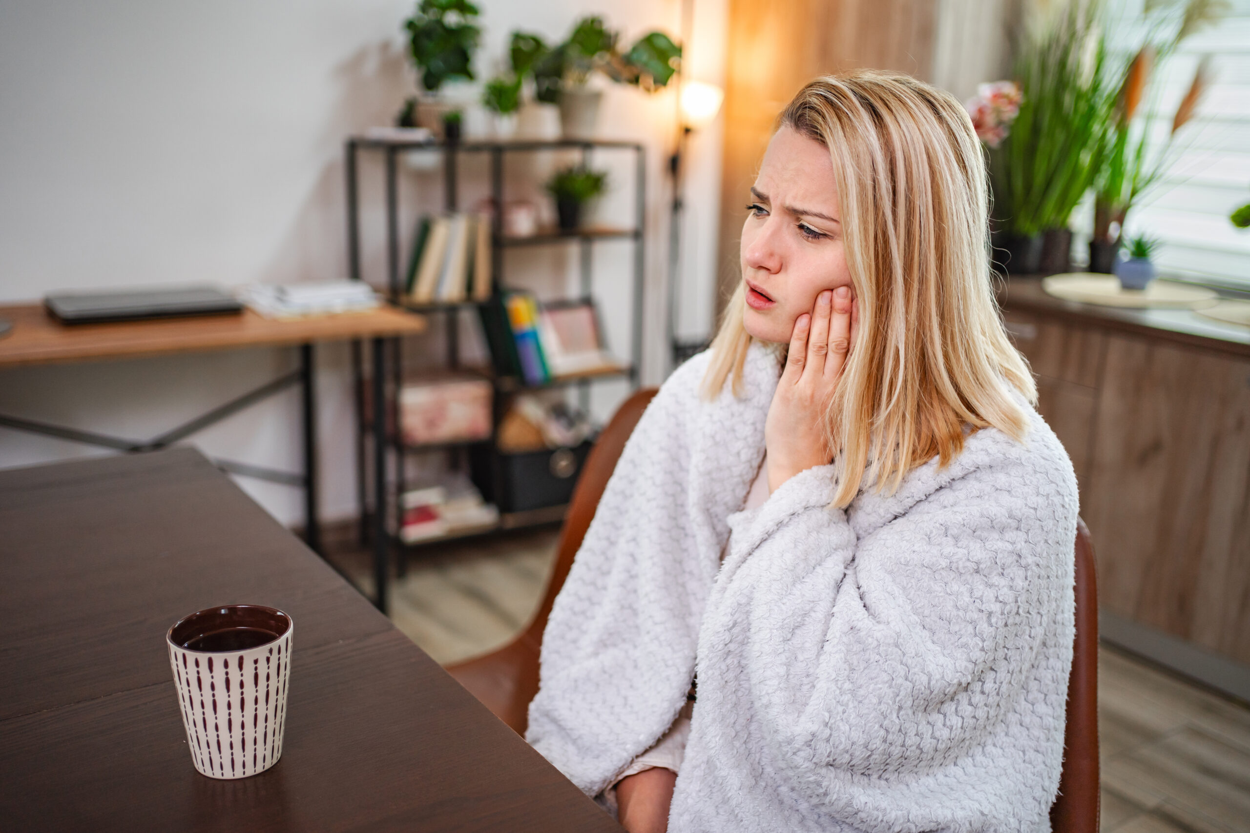 Feeling discomfort while sitting at a cozy table indoors Woman experiencing tooth pain while sitting at home with a cup of coffee