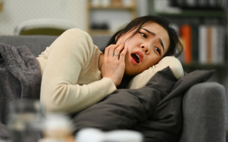 Woman lying on a couch holding her jaw in pain Woman lying on a couch holding her jaw in pain due to a sudden toothache at home
