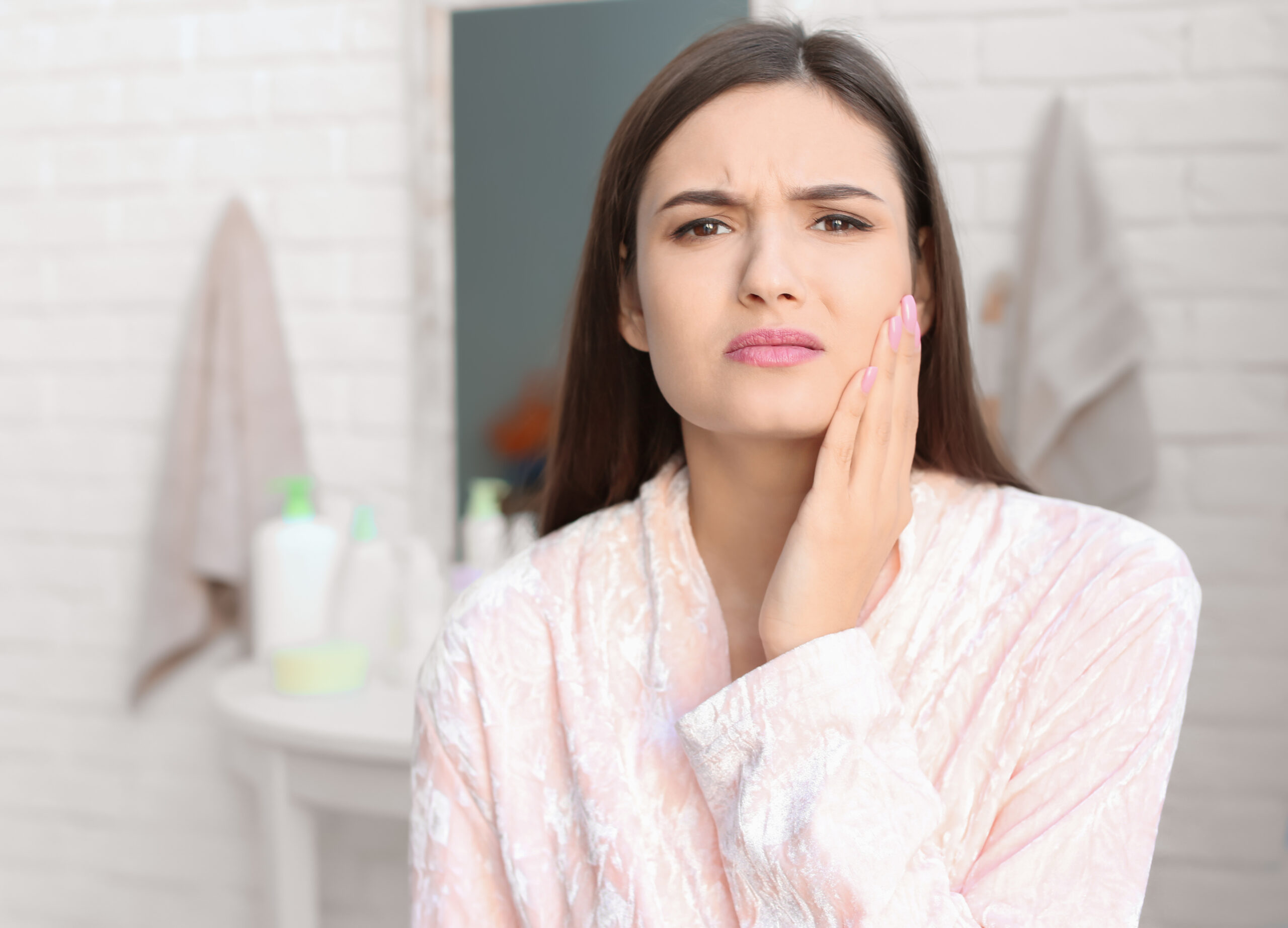Young woman with sensitive teeth in bathroom Woman holding her cheek with tooth pain at night while standing in a bathroom, illustrating severe nighttime toothache discomfort.