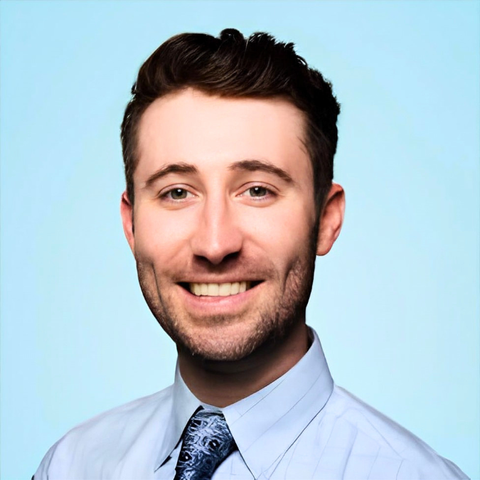 Professional headshot of smiling male dentist wearing light blue shirt and navy tie against blue background.
