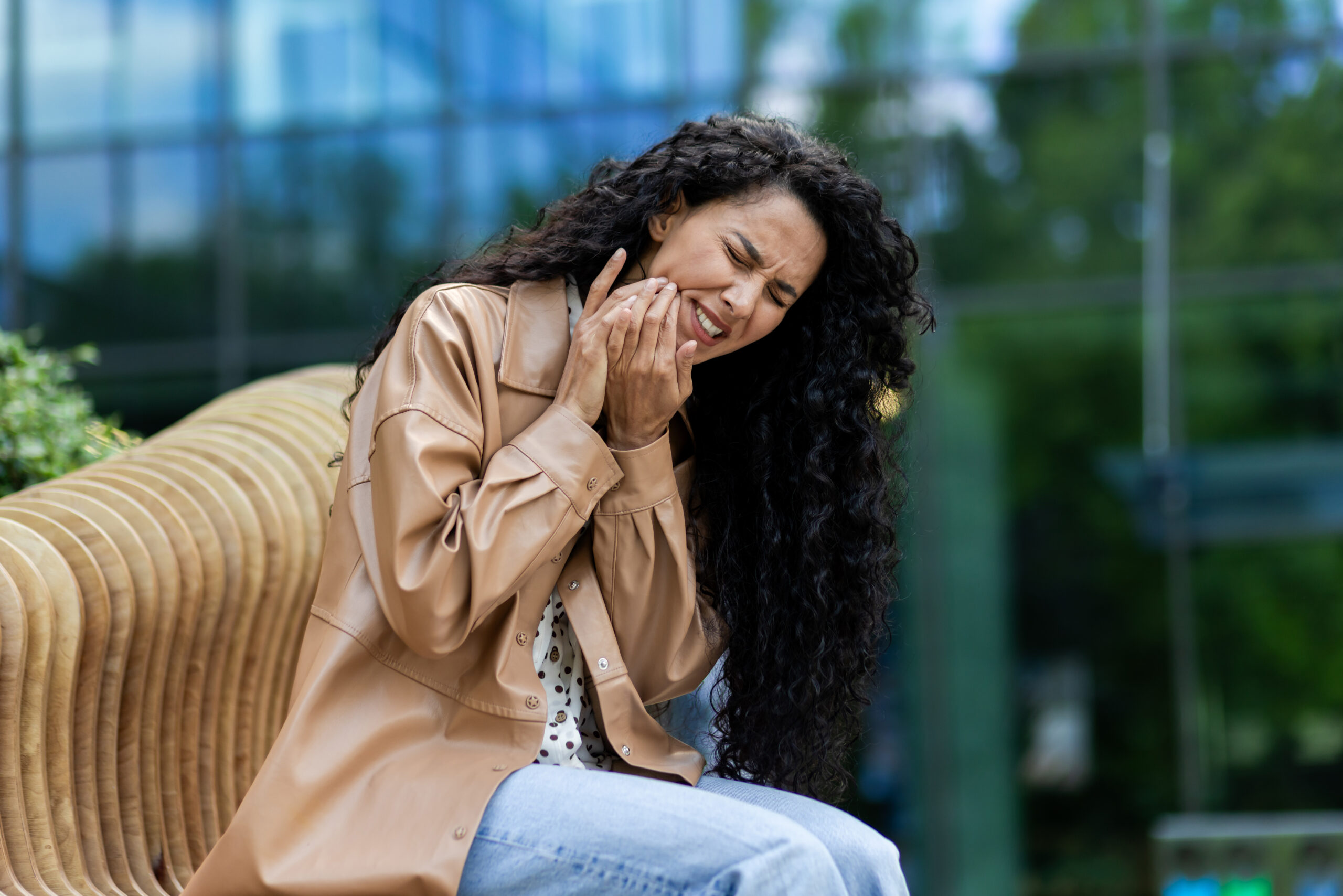 Frustrated caucasian woman holding cheek area and feeling pain in teeth or jaw while sitting on bench outdoors with buildings and greenery on background. Concept of ache and suffering Woman sitting outdoors holding her cheek in pain, showing discomfort from a sudden toothache or dental emergency