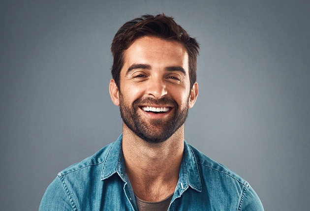 Smiling male dentist in blue shirt seated in modern dental office treatment room.